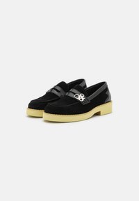 Schwarze Wildleder-Loafers mit einem glänzenden schwarzen Riemen und silbernem Metalldetail, ausgestattet mit einer gelben Plateausohle und gesteppten Verzierungen.