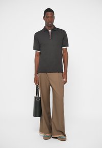 Άνδρας που στέκεται, φορώντας μαύρο πόλο, φαρδιά καφέ παντελόνια, μπλε παπούτσια, κρατώντας μαύρη κυλινδρική τσάντα μπροστά από λευκό φόντο.