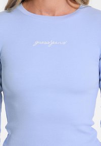 Vaaleansininen, ribbattua pitkähihaista puseroa, jossa on pyöreä pääntie. Rintamuksessa on kirjailtuna kursiivilla "guessjeans".