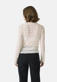 Helles, strukturiertes Longsleeve-Oberteil in Creme mit floralen Mustern. Verfügt über ein gerafftes Design und eine weiche Oberfläche, kombiniert mit schwarzen Hosen.