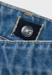 Modrá džínovina s viditelným opotřebením, černý pásek na pase a průhledný knoflík se čtyřmi dírkami, sešitý světlým nitěním.