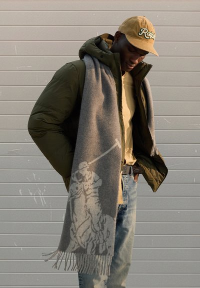 Grå ullskjerf med frynser, med et kontrasterende mønster av en polospiller. Brukes over en grønn dunjakke og beige skjorte.