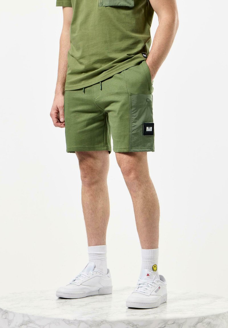 Olivengrønne shorts med en cargo-lomme, snor i livet og et patchmærke på siden. Bæret med en matchende grøn skjorte og hvide sneakers.