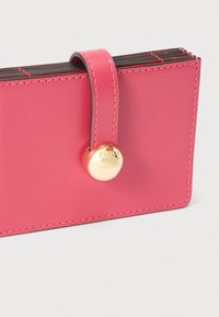 Ružová kožená peňaženka so zlatým gombíkovým zapínaním a prešitými okrajmi. Má zaoblený pásik na vrchu pre bezpečné upevnenie.