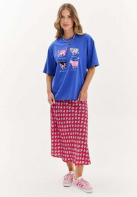 Modré oversized tričko s grafikou krav, doplněné středně dlouhou červenou sukní s modrým vzorem srdcí; vše zakončeno růžovými teniskami.