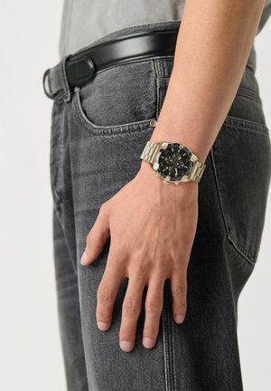 Hand rust zijwaarts met een gouden metalen polshorloge met zwarte wijzerplaat, gecombineerd met een zwarte riem en donkergrijze spijkerbroek.