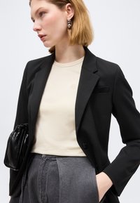 Γυναίκα με κοντά ξανθά μαλλιά φοράει μαύρο σακάκι πάνω από μπεζ τοπ, γκρι ψηλόμεσο παντελόνι, ασημένια κρίκους σκουλαρίκια και κρατά μια μικρή μαύρη δερμάτινη τσάντα.