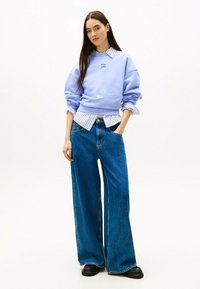Felpa viola chiaro con spalle scese, indossata sopra una camicia a righe blu e bianche. Jeans blu a gamba larga, scarpe chunky nere.