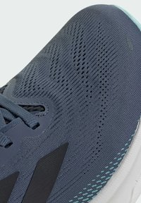 Scarpa sportiva grigio scuro con tomaia in rete traspirante, superficie testurizzata, dettagli neri e suola bianca. Presenta un design con lacci.