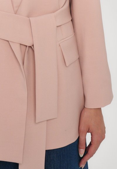 Blush pink wrap coat with a tied belt and side pocket, featuring long sleeves and a smooth, structured fabric. Jeans visible at the bottom.