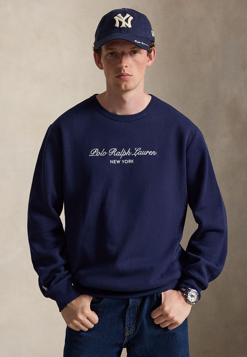 Νέος άνδρας φοράει ένα μπλε σκούρο φούτερ Polo Ralph Lauren, σκούρο τζιν, μπλε σκουρό καπέλο New York Yankees και ένα ρολόι χειρός με σκούρο λουρί.