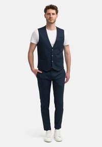 Chaleco azul marino con escote en V, seis botones y dos bolsillos frontales, combinado con pantalones a juego y zapatillas blancas. Tejido texturizado.