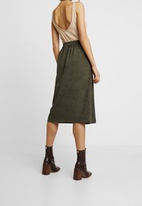 Olivengrøn midi-nederdel med elastisk talje, parret med en beige ribbet tanktop og brune hælede støvler. Glat tekstur og enkelt design.