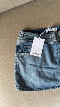Gonna in denim azzurro chiaro con cintura e tasca frontale, adagiata su un tessuto beige, con il tag del marchio Pull&Bear bianco attaccato.