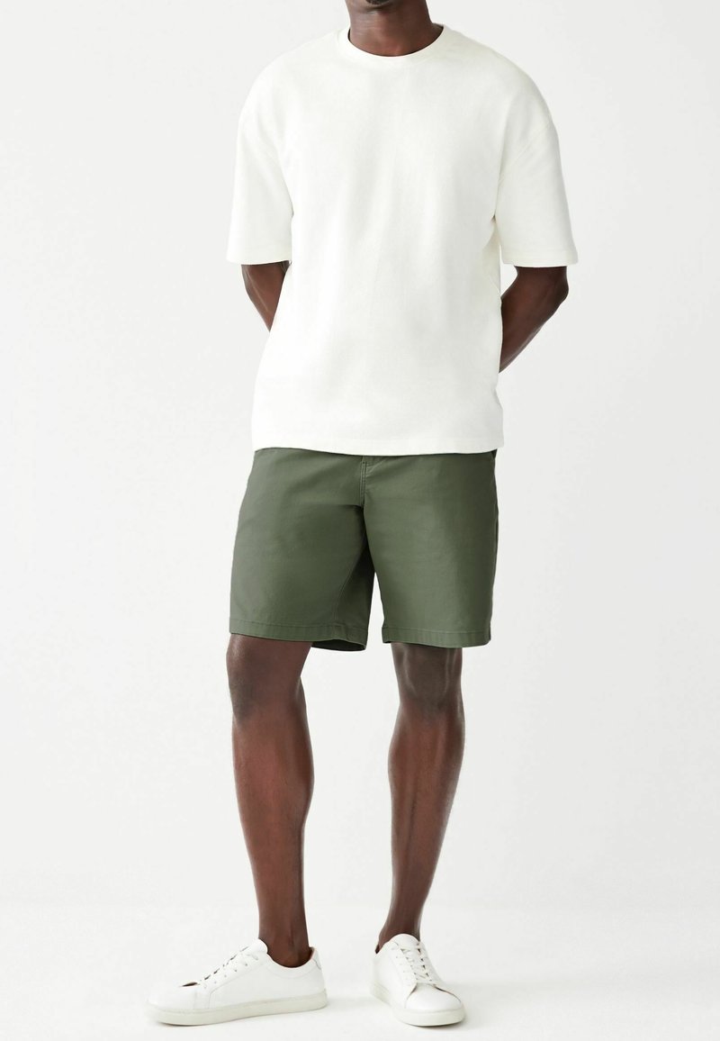Homme portant une chemise blanche à manches courtes simple, un short vert olive et des baskets blanches, debout les mains dans le dos.