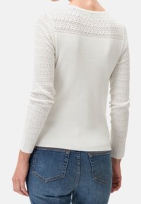 Top bianco a maniche lunghe con corpo a costine e maniche in pizzo testurizzato caratterizzate da motivi geometrici. Il retro presenta una finitura liscia.