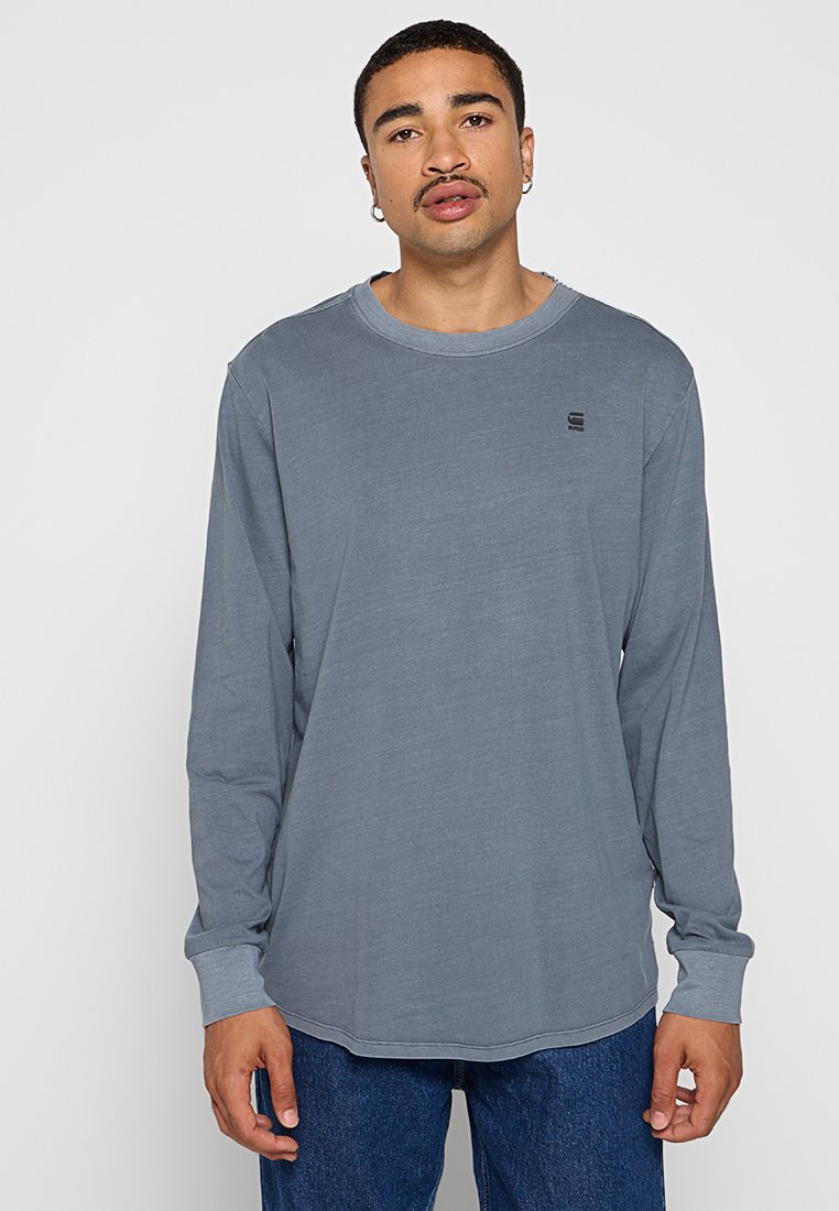 Graues langärmeliges T-Shirt aus einem weichen, leichten Stoff mit Rundhalsausschnitt und dezentem Logo-Akzent auf der Brust. Kombiniert mit blauen Jeans.