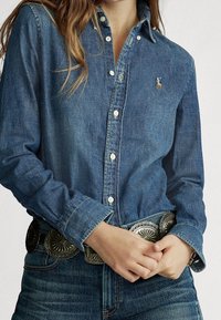 Blå denimskjorta med knappar, krage och långa ärmar, med vita knappar och en liten broderad logotyp på bröstet. Silverfärgad dekorativt bälte.