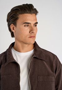 Chaqueta de pana marrón con cremallera y un bolsillo en el pecho, llevada sobre una camiseta blanca. Textura suave y diseño de cuello ajustado.