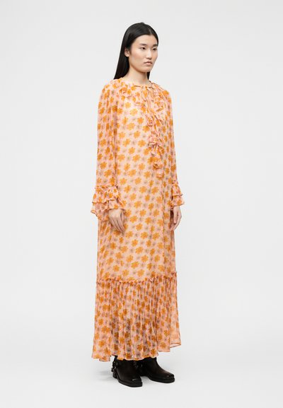 Γυναίκα με μακριά σκούρα μαλλιά, που φοράει μακρύ διαφανές φόρεμα ροδακινί με πορτοκαλί ανθισμένο μοτίβο, βολάν και μαύρα μποτάκια αστραγάλου, στέκεται μπροστά σε λευκό φόντο.