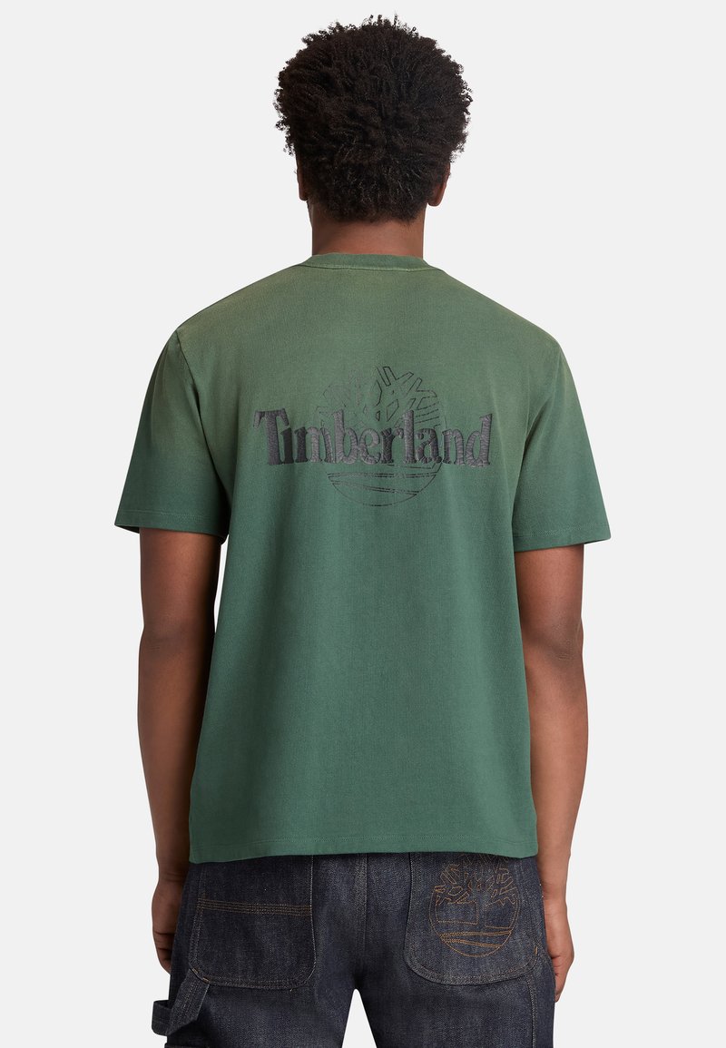 Personne portant un t-shirt Timberland vert avec le logo et un texte au dos, associé à un pantalon en denim foncé avec un logo cousu.