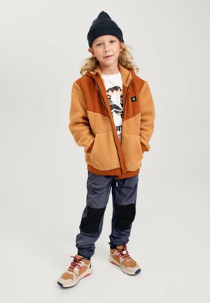 Veste en polaire marron et beige avec zip, pantalon de sport gris et baskets beiges avec accents rouges. L'enfant porte un bonnet noir et un t-shirt graphique.
