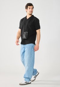 Camisa negra de manga corta con textura, pantalones vaqueros sueltos azul claro y bolso cruzado negro; el modelo lleva zapatillas blancas con rayas negras.