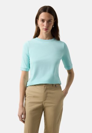 Kobieta z długimi brązowymi włosami nosząca srebrne kolczyki koła, jasnoniebieską bluzkę z krótkim rękawem i beżowe spodnie, z jedną ręką w kieszeni.