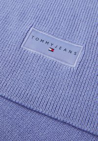 Suéter de punto de color púrpura claro con un patrón texturizado. Presenta una etiqueta rectangular con "TOMMY JEANS" y un logotipo en azul, rojo y blanco.