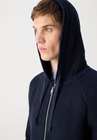 Felpa blu navy con zip, in maglia testurizzata, grande cappuccio e coulisse. Presenta una zip argentata e maniche raglan.