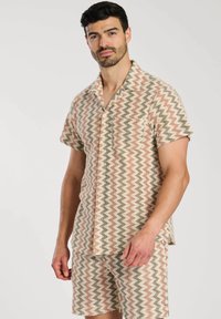 Conjunto de camiseta de manga corta y pantalones cortos con un patrón en zigzag. Los colores incluyen crema, durazno y verde oscuro. Tela ligera y detalle de botones.