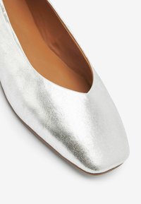 Silverfärgade metalliska ballerinaskor med kvadratig tå och slät yta. Inuti finns en tanfärgad sula. Lätta och minimalistiska i designen.