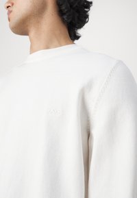 Vitt crewneck sweatshirt i mjukt tyg, med en diskret logotyp broderad på vänster bröst och texturerad söm på ärmarna.