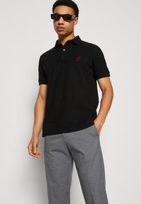 Schwarzes Kurzarm-Polohemd mit Kragen, das mit einem roten Logo-Detail auf der Brust versehen ist. Das Model kombiniert es mit grauen, strukturierten Hosen.