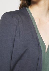 Tmavě šedá bunda s hladkou texturou, s kulatým výstřihem přes zelený top, který zvýrazňuje vrstvený design látky.
