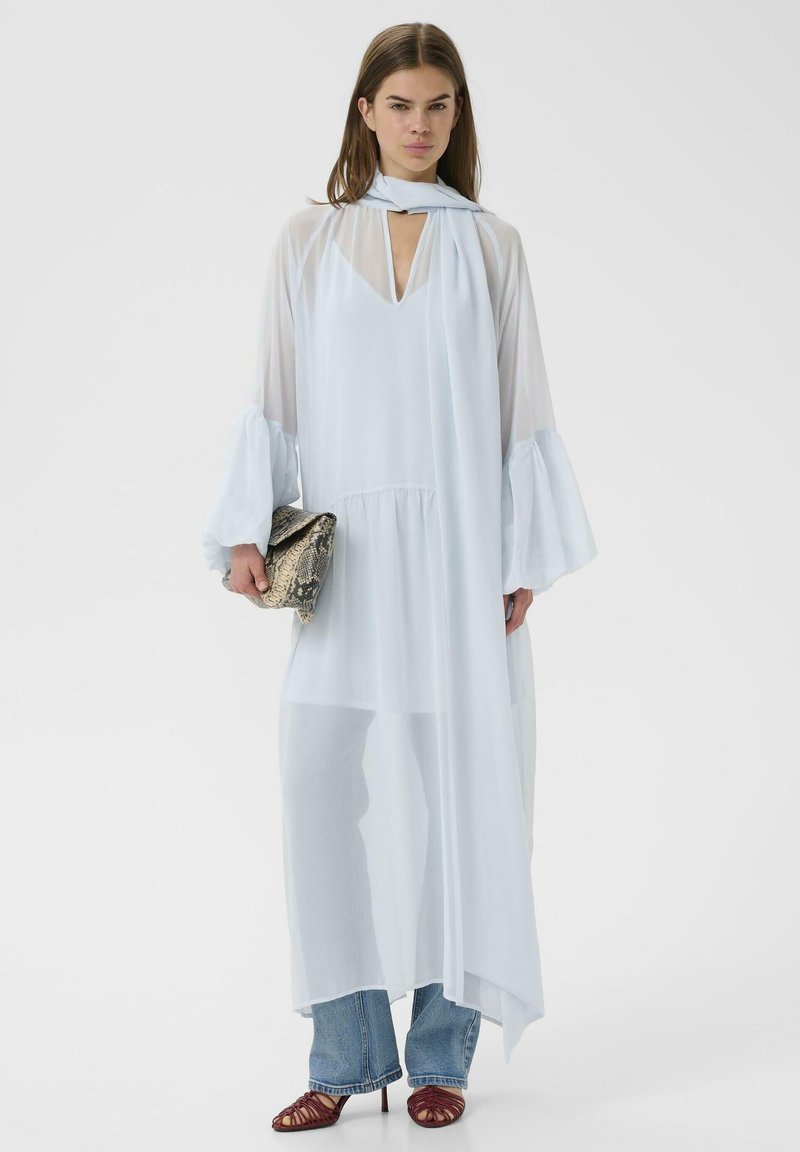 Femme portant une longue robe bleu pâle en tissu transparent avec des manches évasées, un foulard en détail, un jean bleu, des talons rouges à lanières, tenant une pochette en peau de serpent.