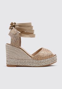 Sandalias de cuña con base de yute trenzado, tiras de tela dorada y diseño de punta abierta. Las tiras se envuelven alrededor del tobillo para asegurar la sujeción.