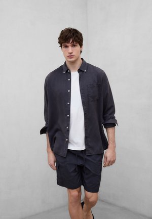 Jonge man die tegen een effen muur staat, gekleed in een zwart overhemd over een wit T-shirt met zwarte korte broek.