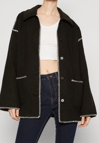 Czarna, oversize kurtka z białymi przeszyciami, posiadająca kołnierz, guziki z przodu oraz dwie łatane kieszenie. Miękka tkanina i luźny krój.