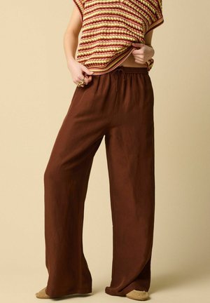 Person trägt lockere braune Hosen mit weitem Bein und Kordelzug in der Taille, hält ein gestreiftes gestricktes Oberteil in Rot-, Gelb- und Cremetönen hoch.