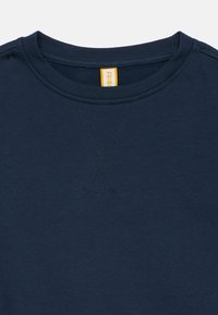 Σκούρο μπλε φούτερ με στρογγυλή λαιμόκοψη, με ραμμένο V-shaped σχέδιο κάτω από το γιακά και ορ�ατή κίτρινη ετικέτα μάρκας μέσα στον λαιμό.