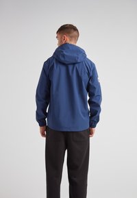 Veste imperméable bleu marine avec une capuche, des poignets élastiques et un détail logo sur la manche. Texture lisse et coupe décontractée. Pantalons noirs.