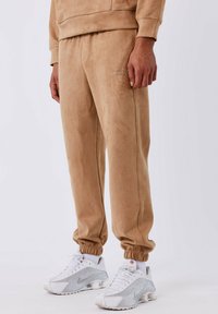 Tan fleece joggers met elastische boorden en subtiele logodetails, gecombineerd met witte sportschoenen en geribde enkelsokken.