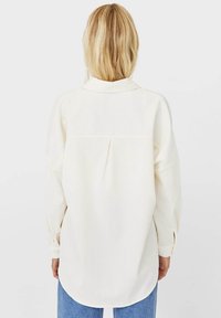 Long-sleeve cream shirt with a collared neck, back pleat detail, and buttoned cuffs, paired with light blue jeans. Soft texture, relaxed fit.