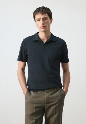 Mladý muž stojaci s rukami v vreckách, oblečený v čiernej polo košeli s krátkym rukávom a hnedých nohaviciach na jednoduchom pozadí.