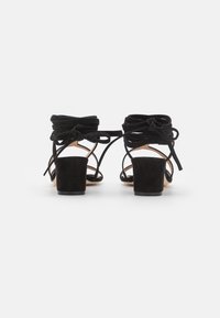 Sandales en daim noir à talons blocs avec des lanières nouées en nœuds autour de la cheville, vues de dos sur fond blanc.