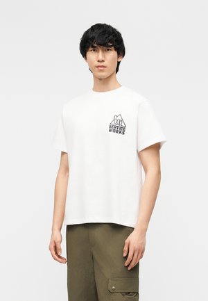 Jonge man die een wit T-shirt draagt met het logo "Service Works" en olijfgroene cargobroek, staand tegen een effen lichte achtergrond.