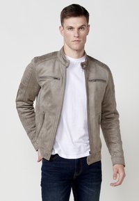 Jachetă din suede gri deschis cu guler în picioare, buzunare cu fermoar și detalii matlasate pe umeri. Pur și simplu peste un tricou alb și blugi negri.