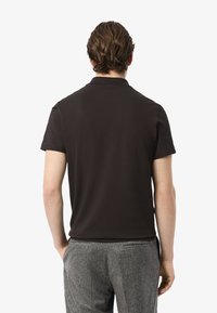 Man med vågigt brunt hår som bär en svart kortärmad polo och grå strukturerade byxor, sedd bakifrån mot en vit bakgrund.