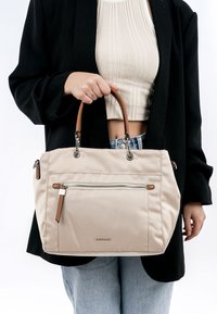 Beige handväska med bruna läderhandtag, som har en framficka med dragkedja och metalliska detaljer. Mjuk tygkvalitet med en strukturerad form.